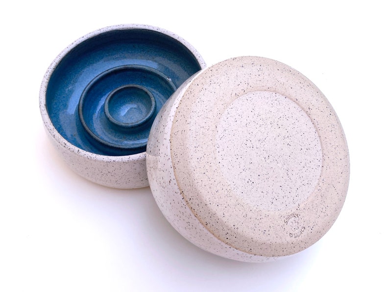 May include: A round ceramic butter keeper with a speckled off-white exterior and a deep blue interior. The base features a central well for water, and the lid is inverted next to it. The text "SUNNY CERAMICS" is stamped on the bottom of the lid.