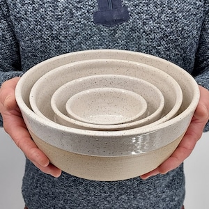 May include: Set of four nesting bowls with speckled white and beige glaze. The bowls are made of ceramic and have a rounded shape.