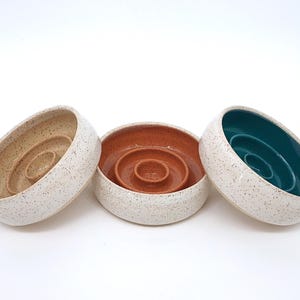 May include: Three round ceramic bowls with a speckled white exterior. Each bowl features a raised circular center pedestal. The interior of the left bowl is beige, the center bowl is terracotta orange, and the right bowl is a deep teal green.