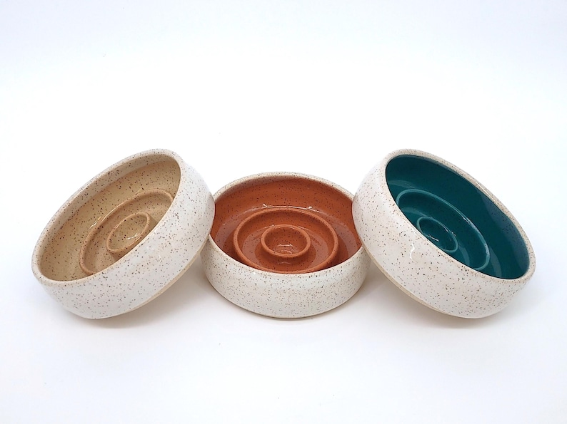 May include: Three round ceramic bowls with a speckled white exterior. Each bowl features a raised circular center pedestal. The interior of the left bowl is beige, the center bowl is terracotta orange, and the right bowl is a deep teal green.