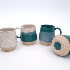 May include: Four ceramic mugs in various shades of teal, speckled white, and beige. The mugs have rounded bodies and large handles. One mug is lying on its side, revealing the beige base. The mugs are suitable for coffee, tea, or other beverages.
