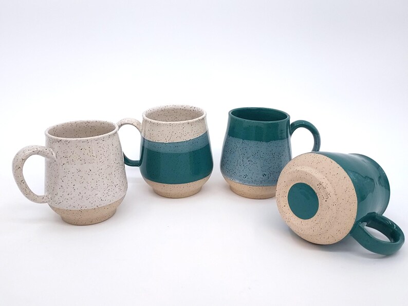 May include: Four ceramic mugs in various shades of teal, speckled white, and beige. The mugs have rounded bodies and large handles. One mug is lying on its side, revealing the beige base. The mugs are suitable for coffee, tea, or other beverages.
