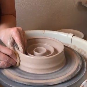 May include: A light brown clay vessel being shaped on a pottery wheel. The circular object features a wide outer rim and a smaller, raised central cup-like section. A small, damp sponge is held against the side of the clay to smooth the surface.