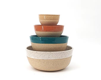 May include: Set of four speckled ceramic bowls in various sizes. The bowls are stacked with the smallest on top. The bowls have a white base with a colored band around the top. The colors are orange, teal, and white.