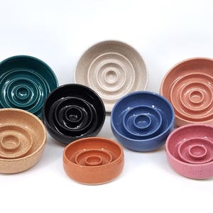 May include: A collection of eight round ceramic bowls in various colors including teal, black, blue, pink, peach, and beige. Each bowl features a distinct concentric ring pattern on the interior base, creating a raised circular center design.
