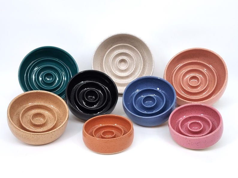 May include: A collection of eight round ceramic bowls in various colors including teal, black, blue, pink, peach, and beige. Each bowl features a distinct concentric ring pattern on the interior base, creating a raised circular center design.