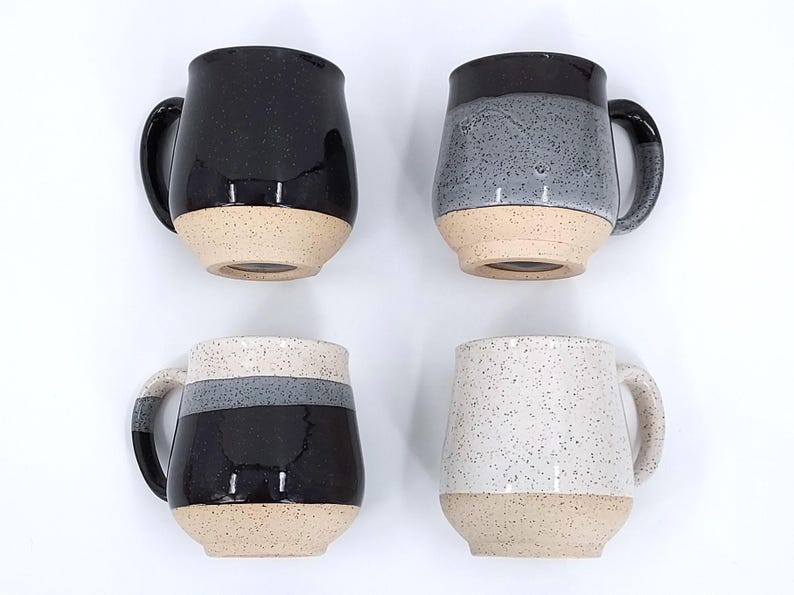 May include: Four ceramic mugs with speckled designs and neutral-colored bases. The mugs feature black, gray, and white speckled patterns. Each mug has a rounded shape with a handle. The mugs are ideal for coffee or tea.