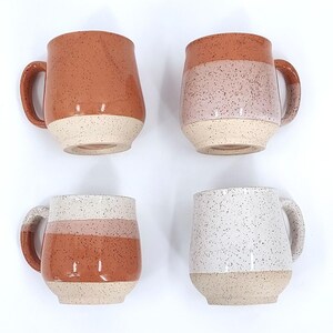 May include: Four ceramic mugs with speckled designs. The mugs feature a rounded shape, large handles, and a two-tone color scheme. Colors include terracotta, pink, and white, with a speckled finish. The base of each mug is a light beige.