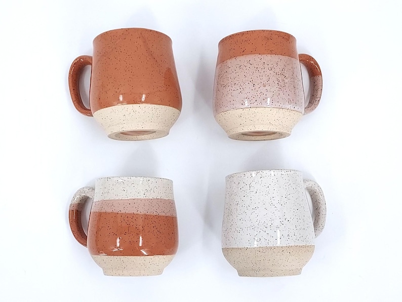 May include: Four ceramic mugs with speckled designs. The mugs feature a rounded shape, large handles, and a two-tone color scheme. Colors include terracotta, pink, and white, with a speckled finish. The base of each mug is a light beige.