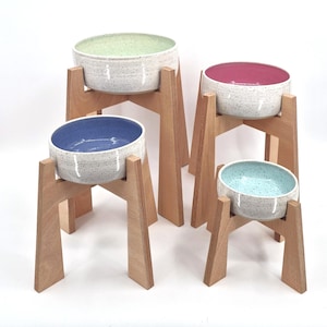 May include: Four elevated pet bowls with wooden stands. Each bowl has a speckled white exterior and a different colored interior: green, pink, blue, and teal. The wooden stands are made of light-colored wood.