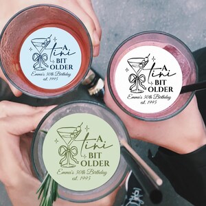 May include: Three cocktails with custom stickers that read "A Tini Bit Older, Emma's 30th Birthday, est. 1995". The drinks are in clear glasses, with a variety of colors, and are being held up by people.