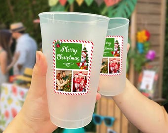 May include: Two clear plastic cups with a Christmas-themed sticker. The sticker has a green background with the words "Merry Christmas" and images of Santa Claus and a family. The cups are held up in front of a blurred outdoor background.
