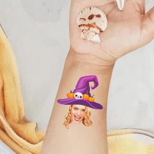 May include: A temporary tattoo featuring a smiling woman's face with a purple witch hat. The hat is decorated with orange pumpkins and a white skull. The tattoo is on a person's arm.