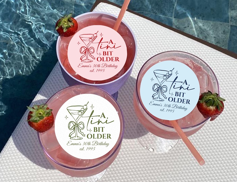 May include: Three cocktails with pink liquid, straws, and strawberry garnishes. Each drink has a circular sticker with a martini glass design and the text "A Tini Bit Older, Emma's 30th Birthday est. 1995". One drink is in a purple cup.