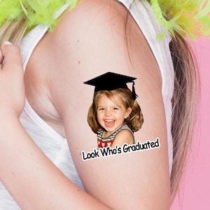 May include: A sticker with the text "Look Who's Graduated" featuring a smiling child wearing a graduation cap. The child is wearing a striped shirt with a heart design. The background is pink.