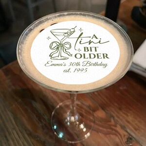 May include: A martini glass filled with a light brown cocktail, topped with a white circular edible image. The image features a martini glass illustration, the text "A Tini Bit Older", and "Emma's 30th Birthday est. 1995" in green.