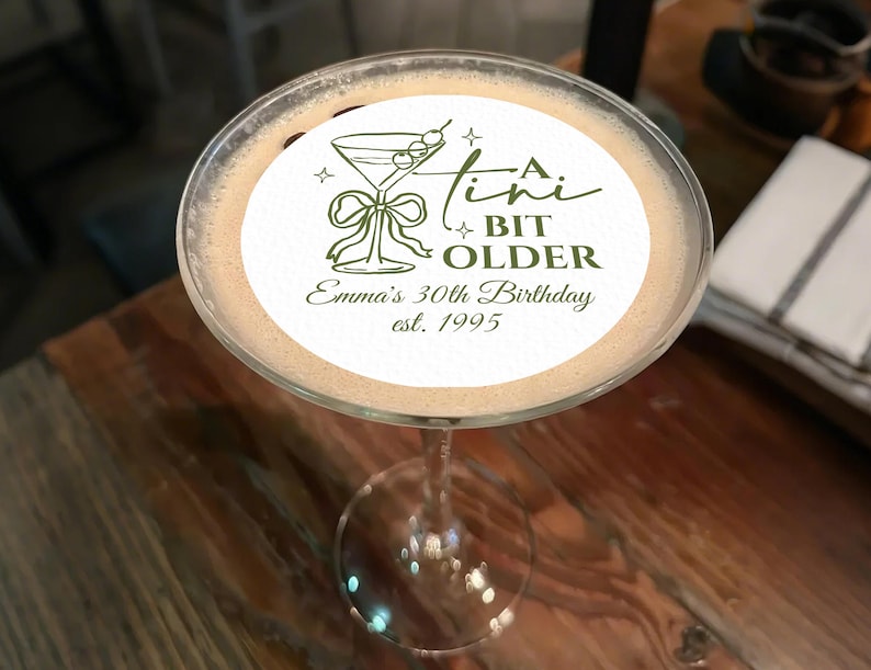 May include: A martini glass filled with a light brown cocktail, topped with a white circular edible image. The image features a martini glass illustration, the text "A Tini Bit Older", and "Emma's 30th Birthday est. 1995" in green.