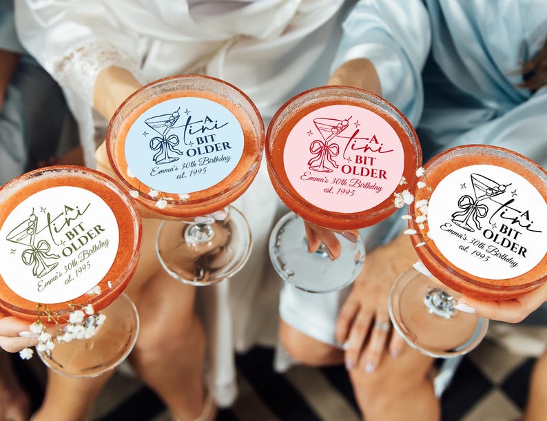 May include: Four cocktails in coupe glasses, each with a circular sticker that reads "A Tini Bit Older, Emma's 30th Birthday, est. 1995." The stickers are in various colors: blue, pink, and white. The drinks are a reddish-orange color.