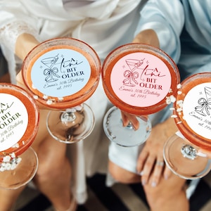 May include: Four cocktails in coupe glasses, each with a circular sticker that reads "A Tini Bit Older, Emma's 30th Birthday, est. 1995." The stickers are in various colors: blue, pink, and white. The cocktails have a reddish-orange hue.