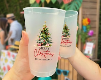 May include: Two frosted plastic cups with a Christmas tree design. The tree is decorated with red and gold ornaments and topped with a gold star. The cups have the words "Merry Christmas" and "The Smith Family" in red script.