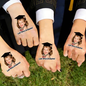 May include: Four hands each displaying a temporary tattoo of a smiling child wearing a graduation cap. The tattoos include the text "Look Who's Graduated" in a white font.