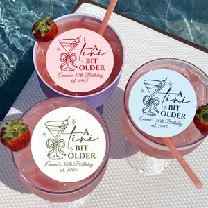 May include: Three cocktails with pink liquid, straws, and strawberry garnishes. Each drink has a circular sticker with a martini glass design and the text "A Tini Bit Older Emma's 30th Birthday est. 1995". One sticker is pink, one is blue, and one is green.
