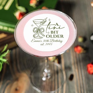 May include: A pink cocktail in a stemmed glass, with a circular label that reads "A Bit Older" and "Emma's 30th Birthday est. 1995". The drink is garnished with a cherry. The background includes books and scattered strawberries.
