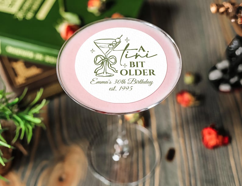 May include: A pink cocktail in a stemmed glass, with a circular label that reads "A Bit Older" and "Emma's 30th Birthday est. 1995". The drink is garnished with a cherry. The background includes books and scattered strawberries.