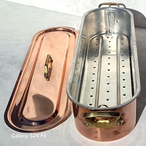 May include: A copper and stainless steel fish poacher with a matching lid. The poacher has a rectangular shape with a perforated interior tray. The lid and handles have a gold finish. The poacher is designed for cooking fish.