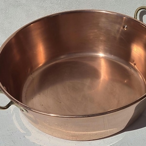 Puede incluir: Una olla de cocina redonda de cobre pulido con un recipiente ancho y poco profundo, y dos asas curvas de latón fijadas a los lados con remaches. Las superficies interior y exterior presentan un acabado de cobre metálico cálido.