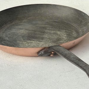 May include: A copper-bottomed frying pan with a dark gray interior and a long, dark metal handle. The pan has a rounded shape and is designed for cooking. The handle has a hole at the end for hanging.