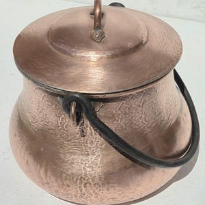 May include: A round, hammered copper pot with a matching lid and a dark, curved metal handle. The pot features a textured, metallic surface with a warm, reddish-brown finish. A small, vertical copper loop handle is centred on the lid.