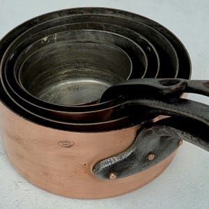 May include: A set of copper pots with iron handles. The pots are nested inside each other, showcasing their varying sizes. The copper exterior has a warm, reddish-brown hue, while the interiors are a contrasting silver color.