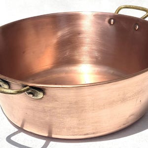 May include: An oval-shaped copper basin with a polished metallic finish and two brass handles attached to the sides with rivets. The interior and exterior surfaces show a warm reddish-brown hue with subtle horizontal brush marks across the metal.
