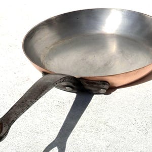 Può includere: Una padella rotonda con esterno in rame lucido e rivestimento interno color argento. La padella presenta un lungo manico in ferro scuro e robusto, fissato lateralmente con due rivetti metallici visibili che si estendono dal corpo circolare.