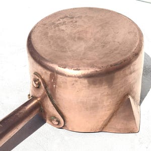 May include: A round copper saucepan featuring a hammered texture on the base and sides. The pan includes a long, straight copper handle attached with two visible rivets and a small triangular pouring spout on the side of the vessel.