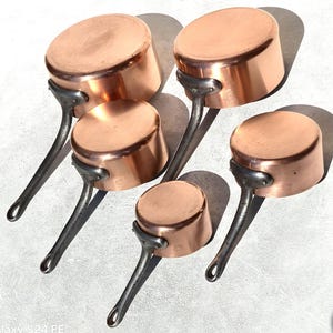 Könnte beinhalten: Fünf Kupfertöpfe mit langen, dunklen Metallgriffen sind auf einer hellen Oberfläche angeordnet. Die Töpfe haben eine glänzende, rotbraune Außenseite und eine abgerundete Form. Die Griffe sind an den Seiten vernietet.