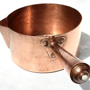 May include: A round copper saucepan featuring a hammered exterior texture, a prominent pouring spout on the side, and a sturdy metal handle attached with a triangular plate. The handle ends in a decorative, tiered knob with a small circular inlay.