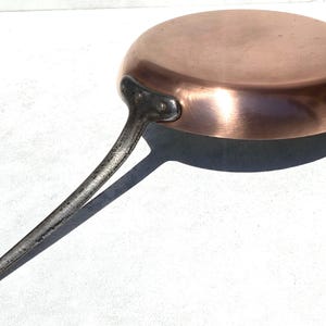 Pode incluir: Uma frigideira redonda de cobre com acabamento metálico polido e um cabo longo de ferro escuro e texturizado. O cabo está fixado de forma segura na lateral da frigideira com dois rebites visíveis. A frigideira tem uma superfície lisa.