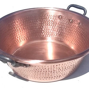 Könnte beinhalten: Eine Kupferschüssel mit gehämmertem Muster und zwei dunklen Metallgriffen. Die Schüssel hat ein breites, flaches Design und eine glänzende, reflektierende Oberfläche. Ideal zum Kochen oder Servieren.