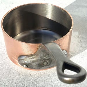 May include: A copper saucepan with a stainless steel interior and a riveted, dark metal handle. The pot has a rounded shape and is designed for cooking. The exterior has a polished copper finish.