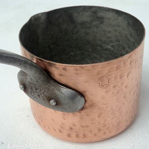 May include: A copper saucepan with a riveted metal handle. The pot has a hammered texture and a dark interior. The handle is attached with two rivets. The pot has a stamp that says "FABRIQUE FRANCE".