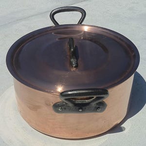 Può includere: Una pentola rotonda in rame con coperchio abbinato. La pentola presenta due manici in metallo scuro e robusto fissati ai lati con rivetti. Anche il coperchio include una maniglia centrale in metallo scuro. La superficie ha una finitura lucida.