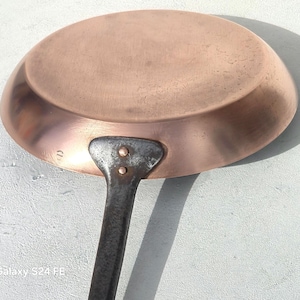 May include: A copper frying pan with a dark metal handle. The pan has a rounded shape and a slightly textured surface. The handle is riveted to the pan. The pan is designed for cooking.