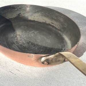 May include: Copper frying pan with a brass handle. The pan has a rounded shape and a dark interior. The handle is attached with rivets. The words "MADE IN FRANCE" are stamped on the side of the pan.