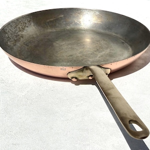 May include: A round copper frying pan with a silver-toned interior and a long, straight metal handle. The exterior side features a copper finish, while the handle is attached with two rivets. The text "MADE IN FRANCE" is stamped near the handle.