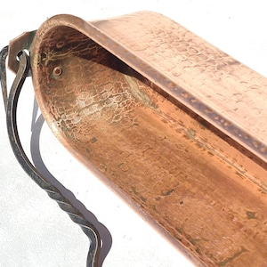 Puede incluir: Un recipiente de cobre martillado de forma alargada, rectangular con esquinas redondeadas. La superficie interior presenta una textura distintiva con hoyuelos. Un asa de metal oscuro y retorcido está unida al lateral con un soporte plano remachado.