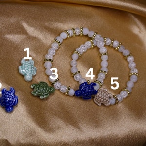 May include: A selection of turtle-themed jewellery. Two bracelets with white and clear beads, gold spacers, and turtle charms. The charms are light blue, dark blue, green, and beige. The background is a gold satin material.