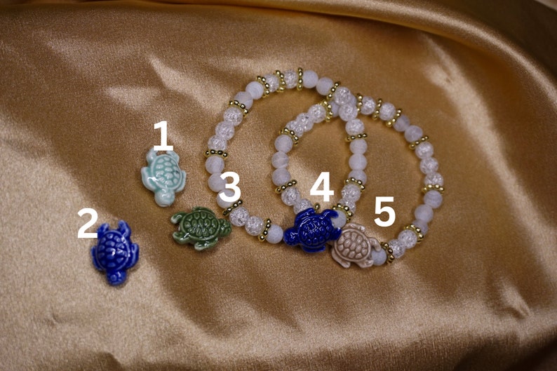 May include: A selection of turtle-themed jewellery. Two bracelets with white and clear beads, gold spacers, and turtle charms. The charms are light blue, dark blue, green, and beige. The background is a gold satin material.
