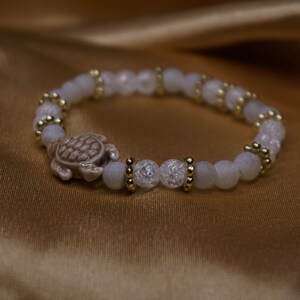 May include: A bracelet with white, crackled beads and gold-coloured accents, featuring a turtle charm. The turtle is light brown. The bracelet is displayed on gold-coloured fabric. The beads are approximately 0.8 cm in diameter.